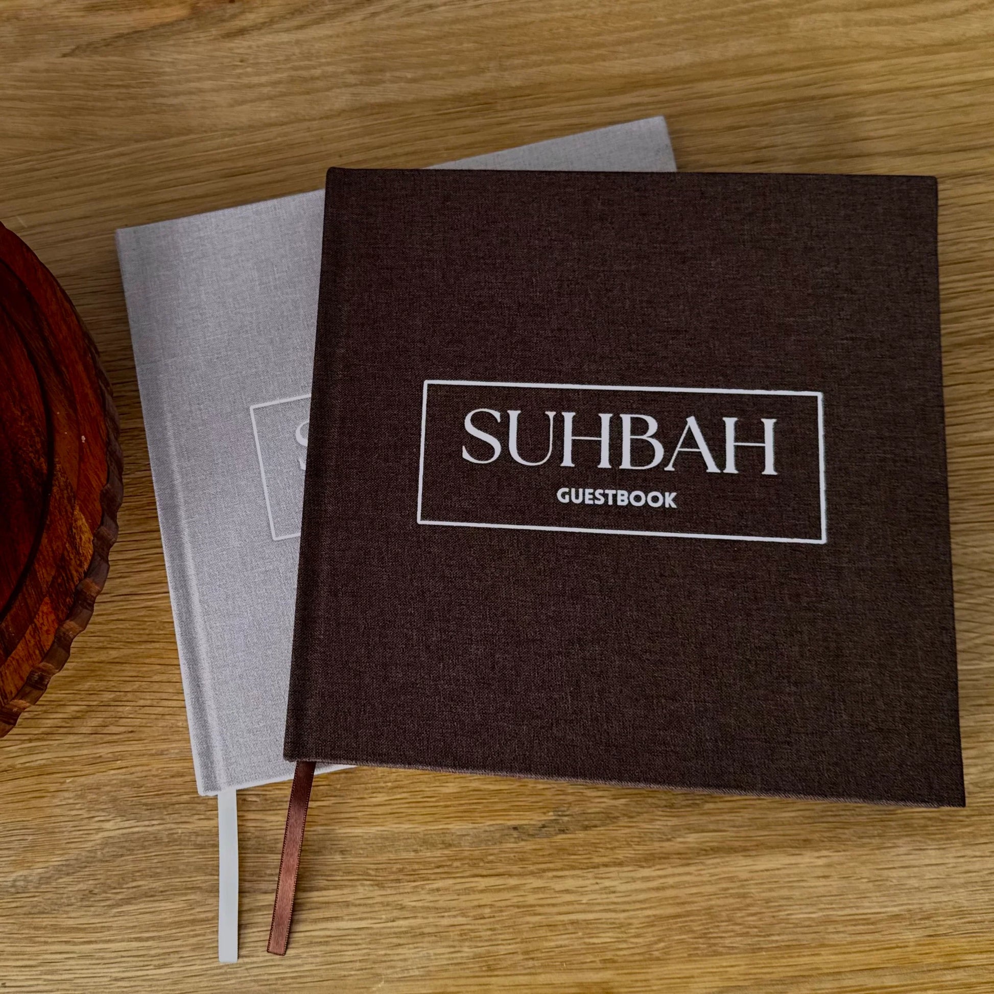 A dark brown linen-textured guestbook with a white printed logo reading 'SUHBAH GUESTBOOK' sits on a light gray guestbook on a wooden tabletop. A brown ribbon bookmark is visible, and part of a dark wooden bowl appears at the left edge.