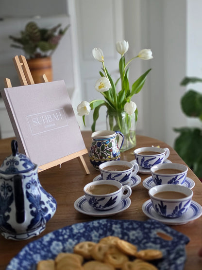 A styled tabletop scene with multiple blue-and-white ceramic teacups and saucers, a matching teapot, a small floral creamer, a plate of cookies, white tulips in a glass vase, and a sign on a wooden easel in a bright indoor setting.