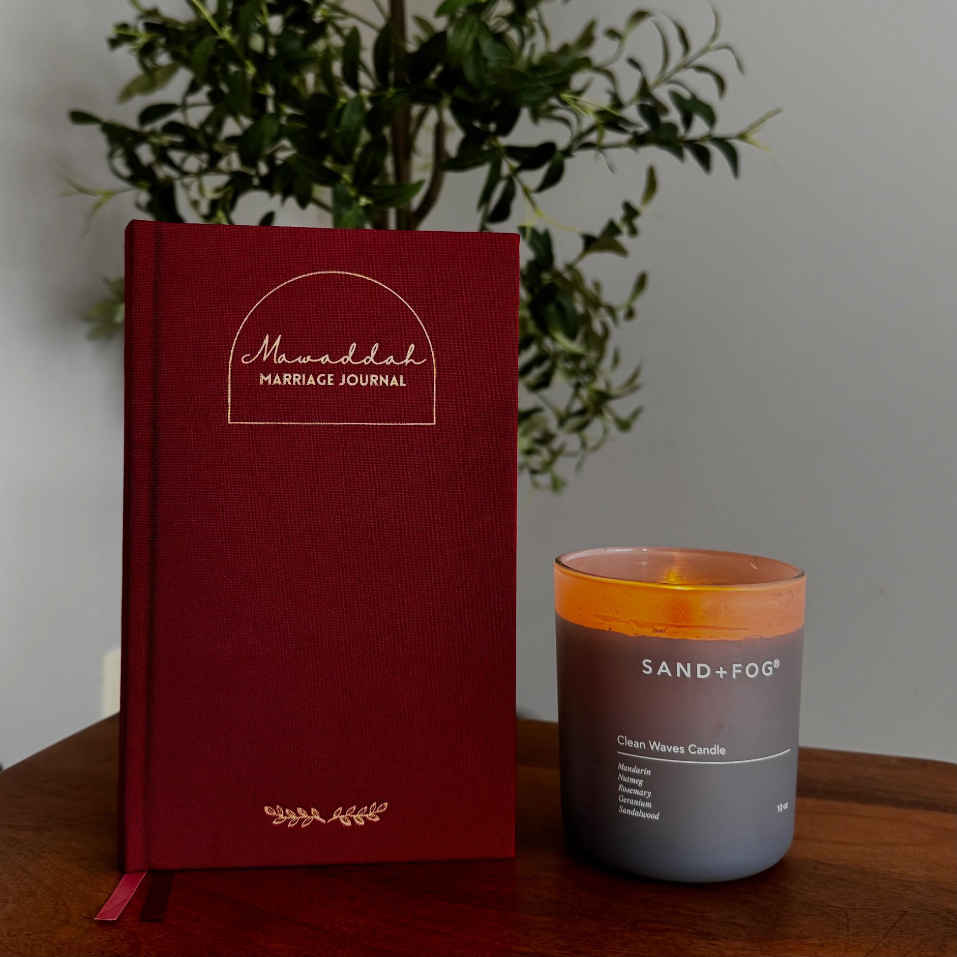 A deep burgundy hardcover marriage journal with gold foil lettering and a ribbon bookmark sits on a wooden table next to a lit Sand + Fog cream wave candle in a gray glass jar, with a green plant in the background.